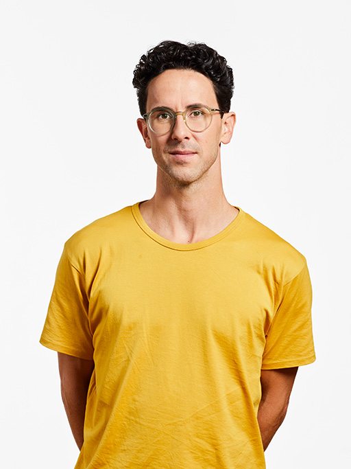 Bild eines Mitarbeiters in gelbem T-Shirt und mit Brille, der seine Arme hinter dem Rücken hält