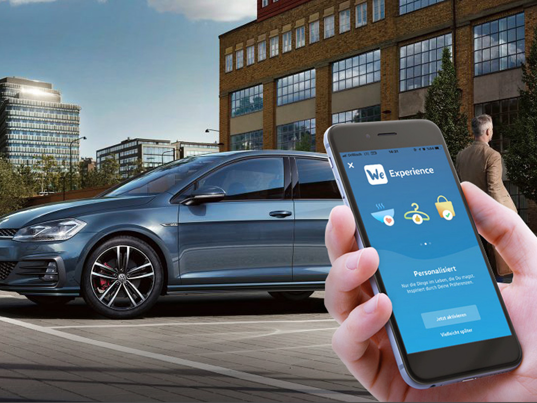 Im Hintergrund erkennt man ein Gebäude und einen blauen VW, im Vordergrund ist ein Smartphone mit der geöffneten VW we App