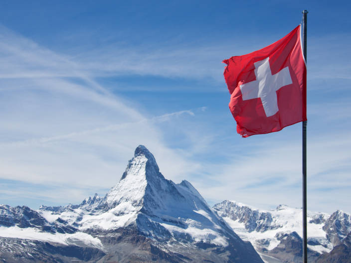 Ein Bild zeigt eine Schweizer Flagge, die über schneebedeckten Bergen weht.
