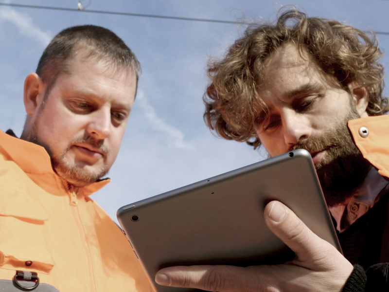 zwei Männer schauen mit orangener Arbeitskleidung auf ein Tablet. Die Kameraperspektive ist von unten