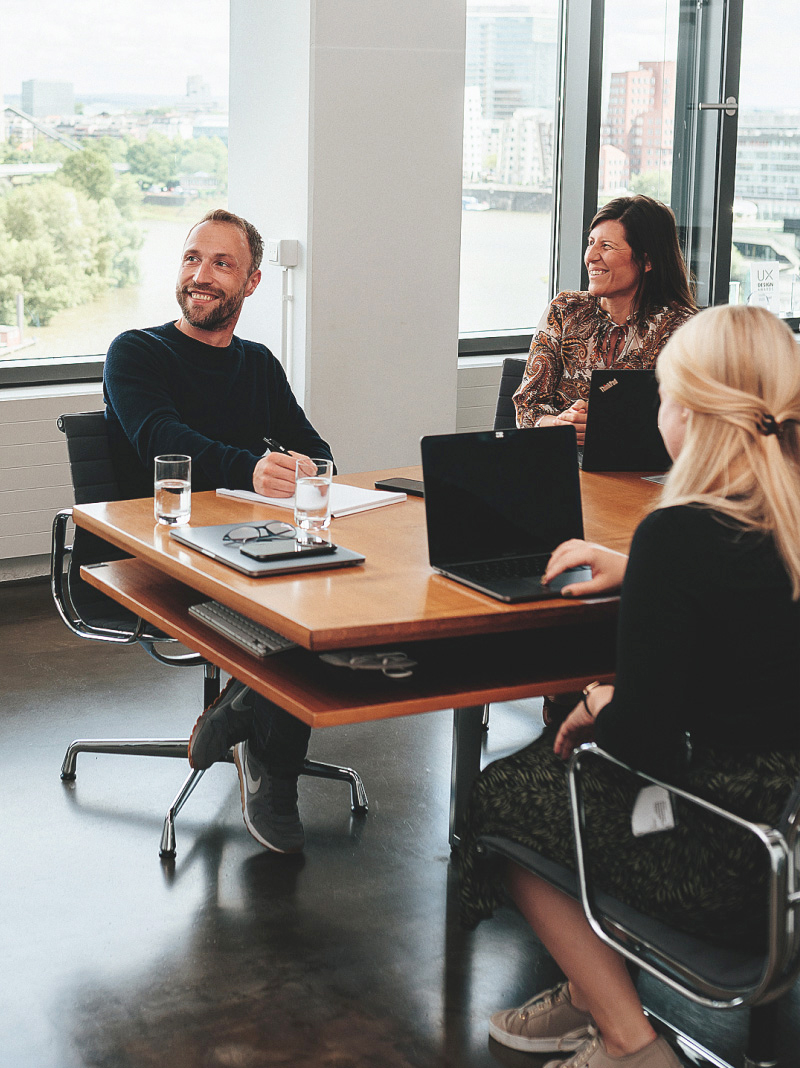 ein Mann sitzt lächelnd mit 2 Arbeitskollegen im Büro bei einem Meeting. Sie schauen zur Seite