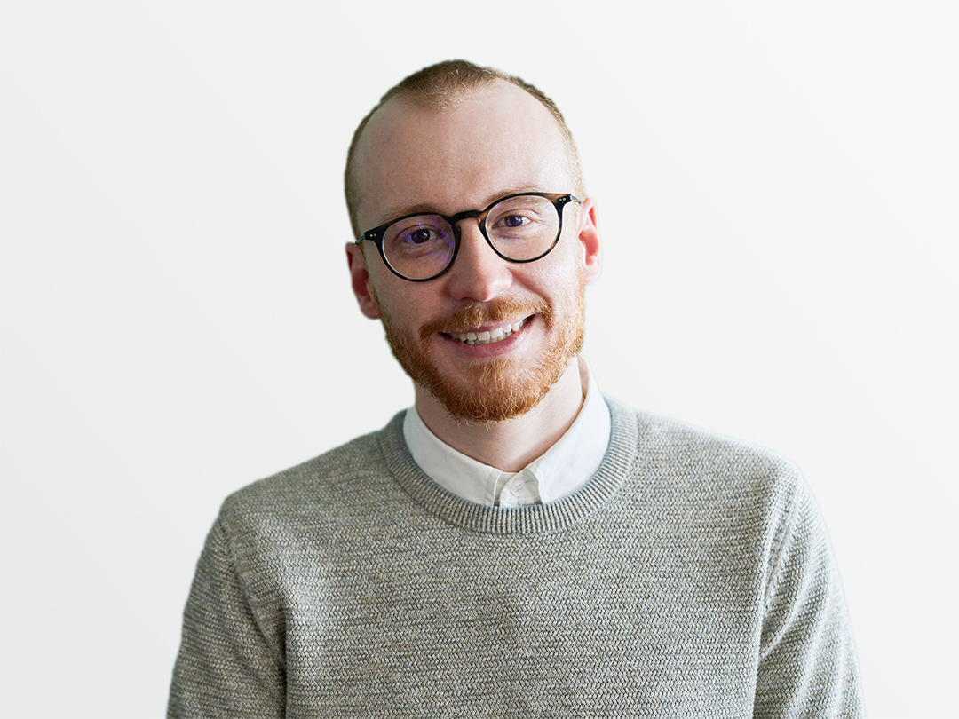 Man with short hair in light grey jumper in front of green liled wall.