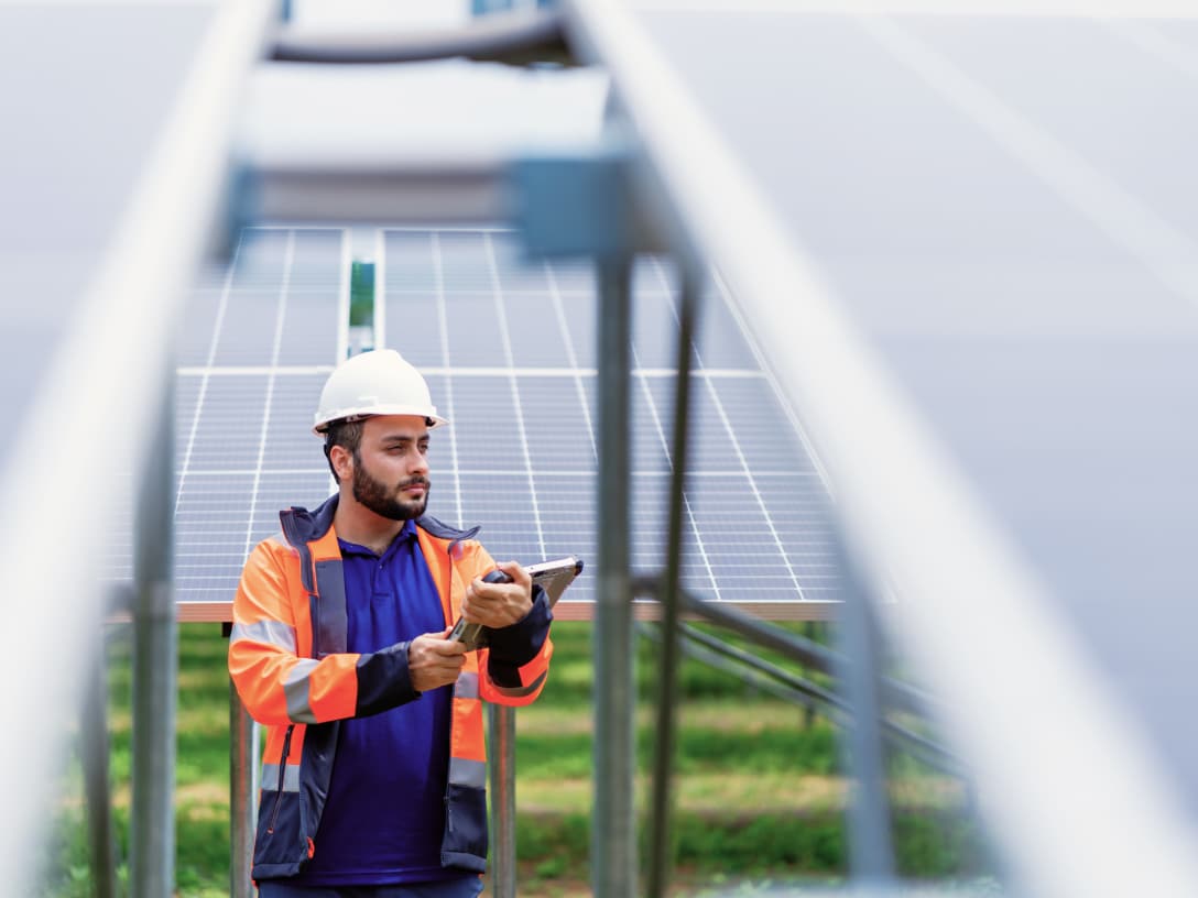 Person mit Helm steht zwischen Photovoltaik-Anlagen, mit Tablet in der Hand