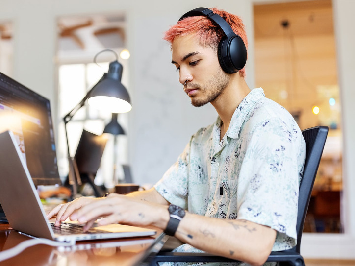 Person with pink hair and headphones working on computer in office environment.