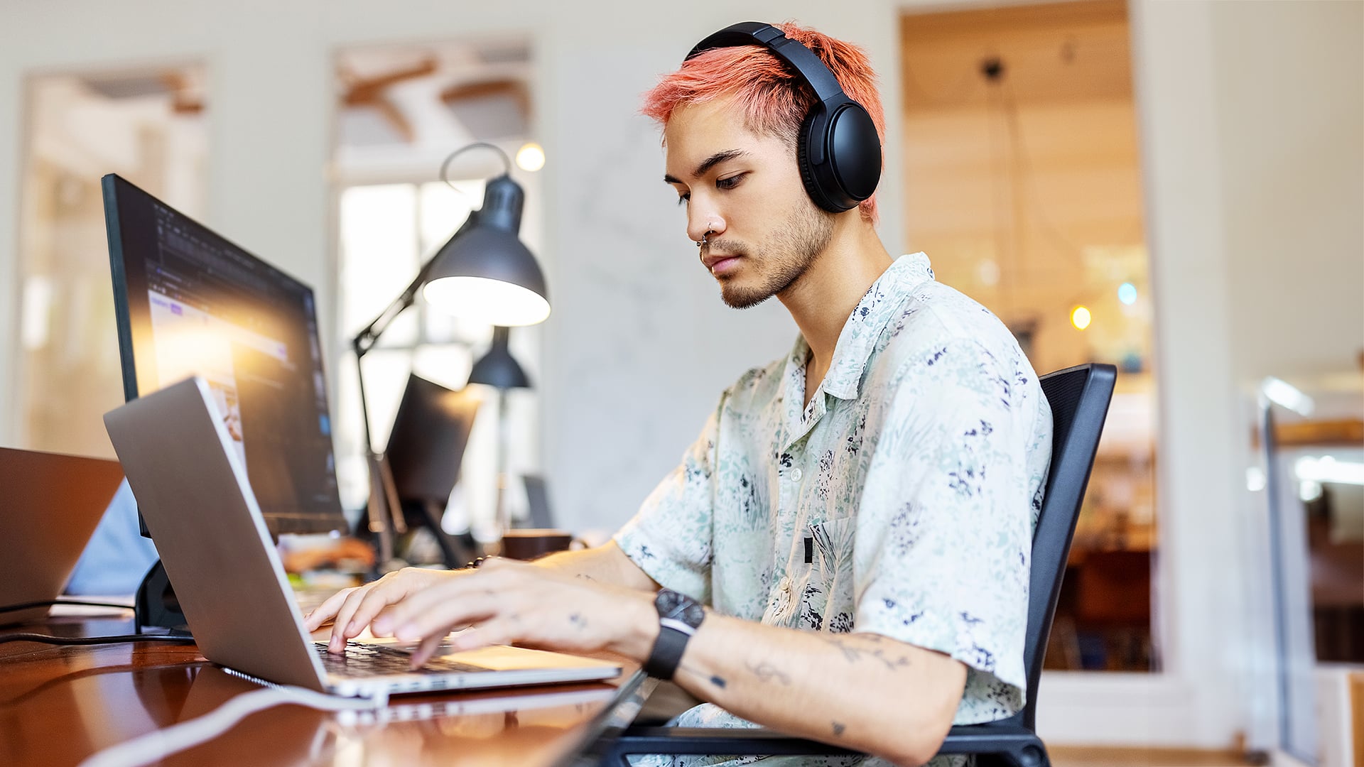 Person with pink hair and headphones working on computer in office environment.