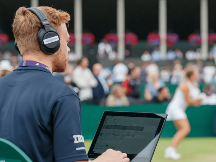 Ein Mann mit Kopfhörern sitzt vor einem Laptop und schaut in Richtung eines Tennisplatzes