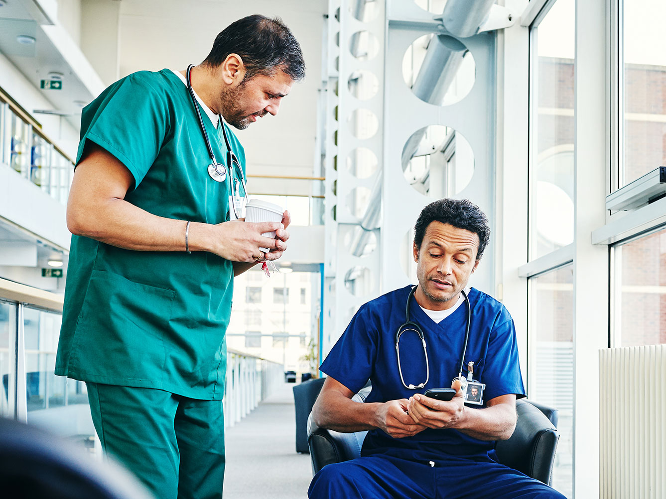 Zwei medizinische Fachkräfte befinden sich in einer medizinischen Einrichtung. Der eine, stehend, trägt einen grünen Kittel und hält eine Kaffeetasse. Der andere, sitzend, trägt einen blauen Kittel und schaut auf ein Smartphone.