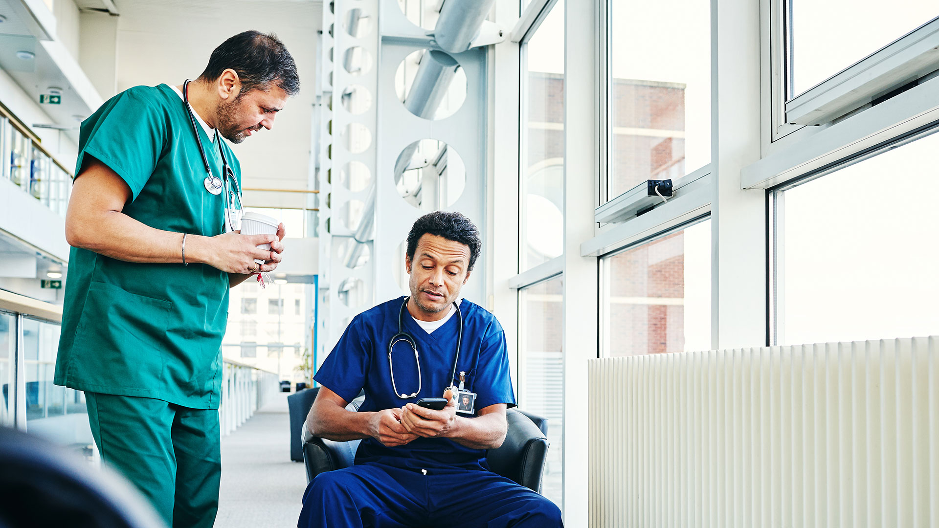 Two healthcare professionals are in a medical facility. One, standing, wears green scrubs while holding a coffee cup. The other, seated, wears blue scrubs and looks at a smartphone.