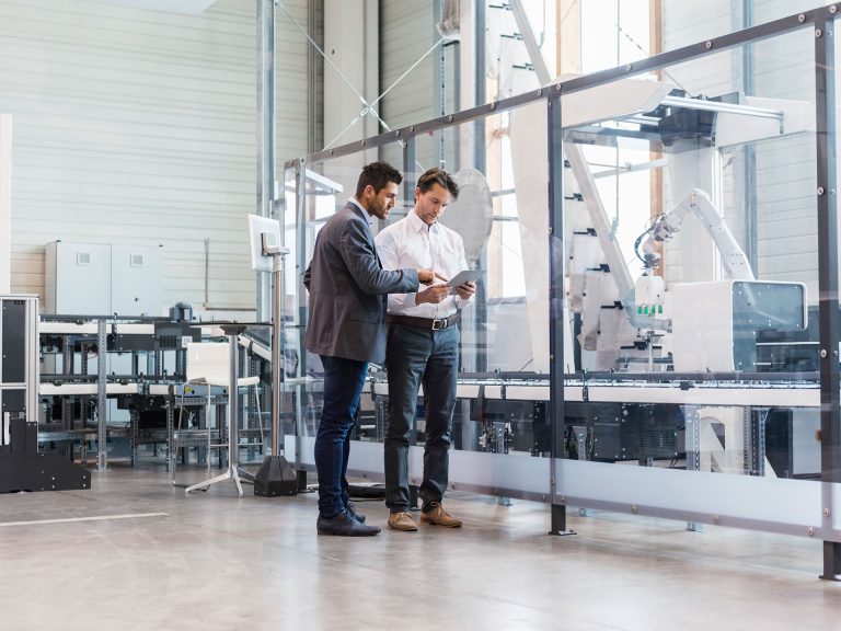 Zwei Mitarbeiter in Anzug stehen in einem Industrielab und schauen auf ein Dokument