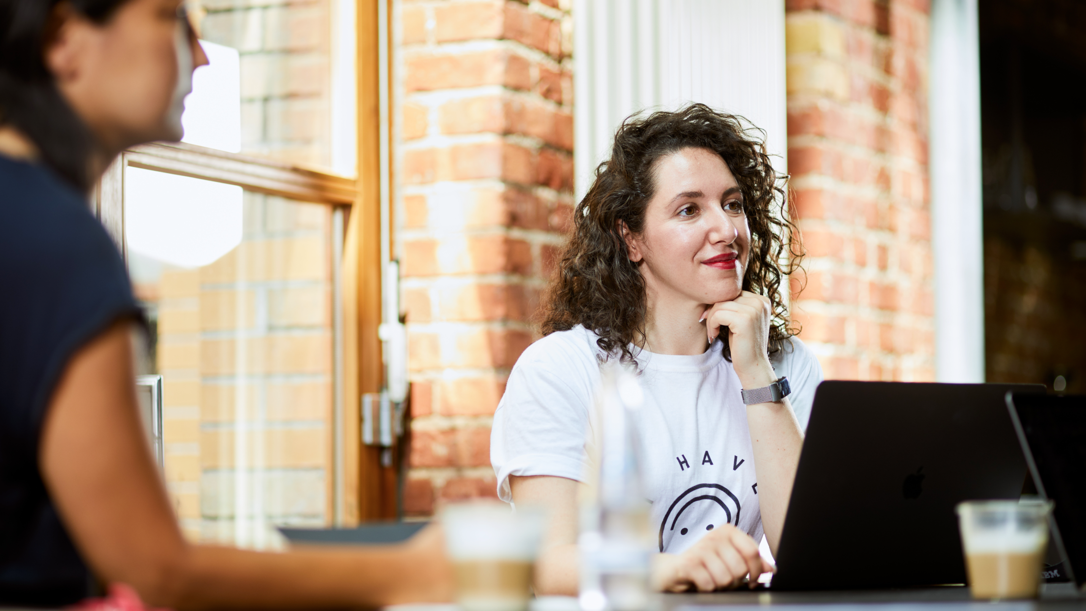 Two people sit at a table with drinks. One, wearing a white t-shirt, smiles while looking at a laptop. The setting has brick walls and large windows.