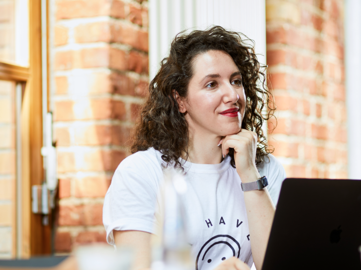 Two people sit at a table with drinks. One, wearing a white t-shirt, smiles while looking at a laptop. The setting has brick walls and large windows.