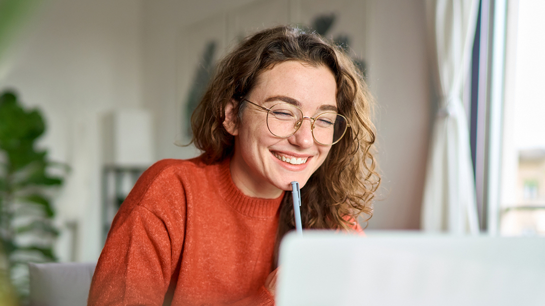Eine Person mit lockigem Haar, die einen orangefarbenen Pullover trägt, hält einen Stift nahe am Mund und schaut auf einen Laptop-Bildschirm. Im Hintergrund sind verschwommene Pflanzen und Möbel zu sehen.