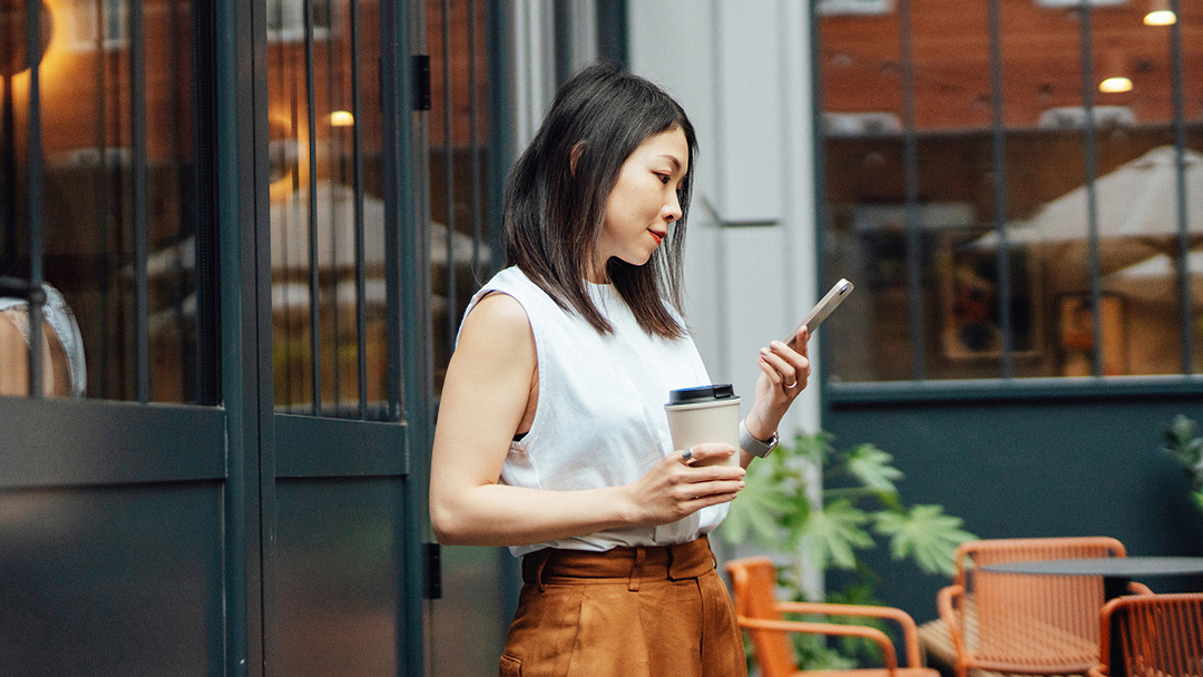 Person steht draußen und hält ein Smartphone und eine Kaffeetasse.