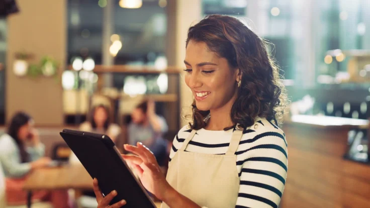 Restaurant worker in apron using tablet in bright café with seated customers