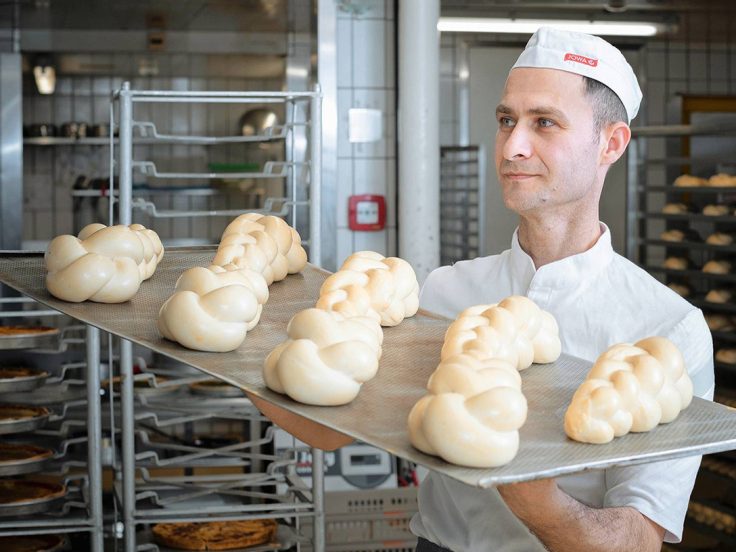 Bäcker in weißer Uniform mit Blech voller Zopfteig in industrieller Bäckereiküche.