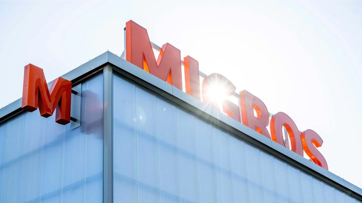 MIGROS-Schriftzug in orange auf einem Gebäude mit hellem Sonnenlicht im Hintergrund.