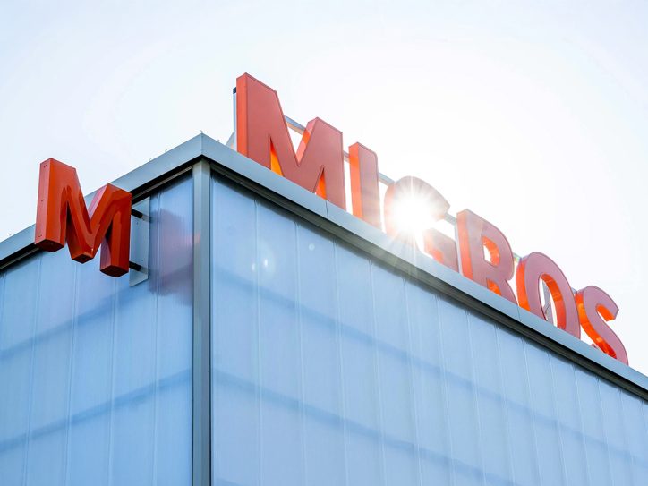 MIGROS-Schriftzug in orange auf einem Gebäude mit hellem Sonnenlicht im Hintergrund.