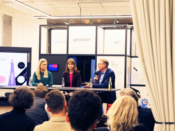 Drei Personen diskutieren auf einer Bühne vor Publikum bei einer Podiumsdiskussion.
