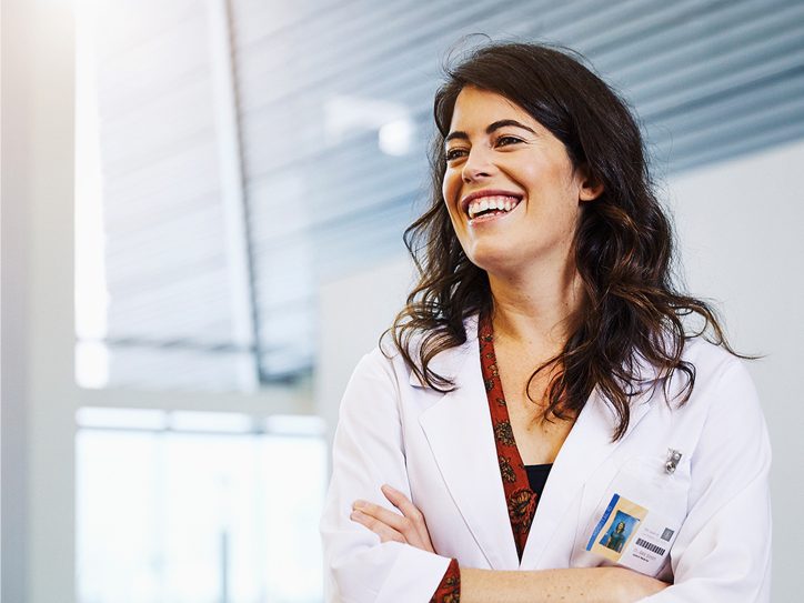 Woman in white lab coat smiling with arms crossed, standing in a modern medical or clinical setting