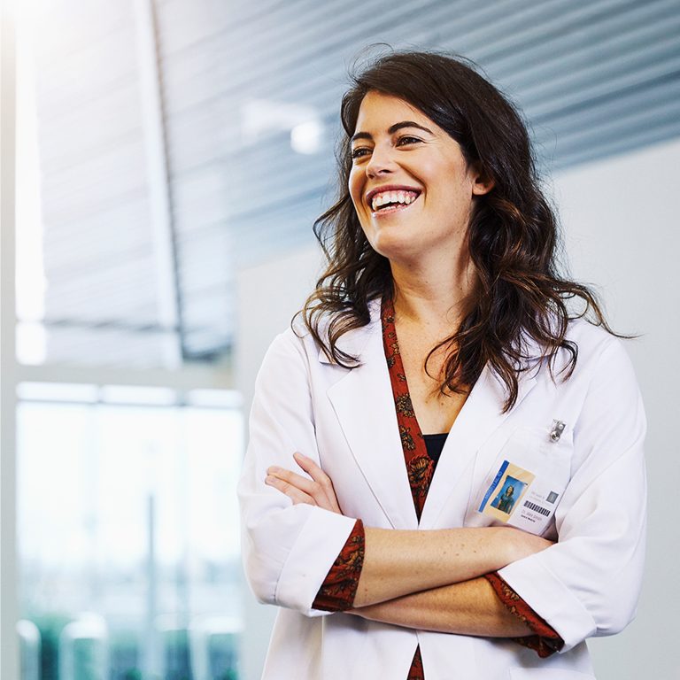 Woman in white lab coat smiling with arms crossed, standing in a modern medical or clinical setting