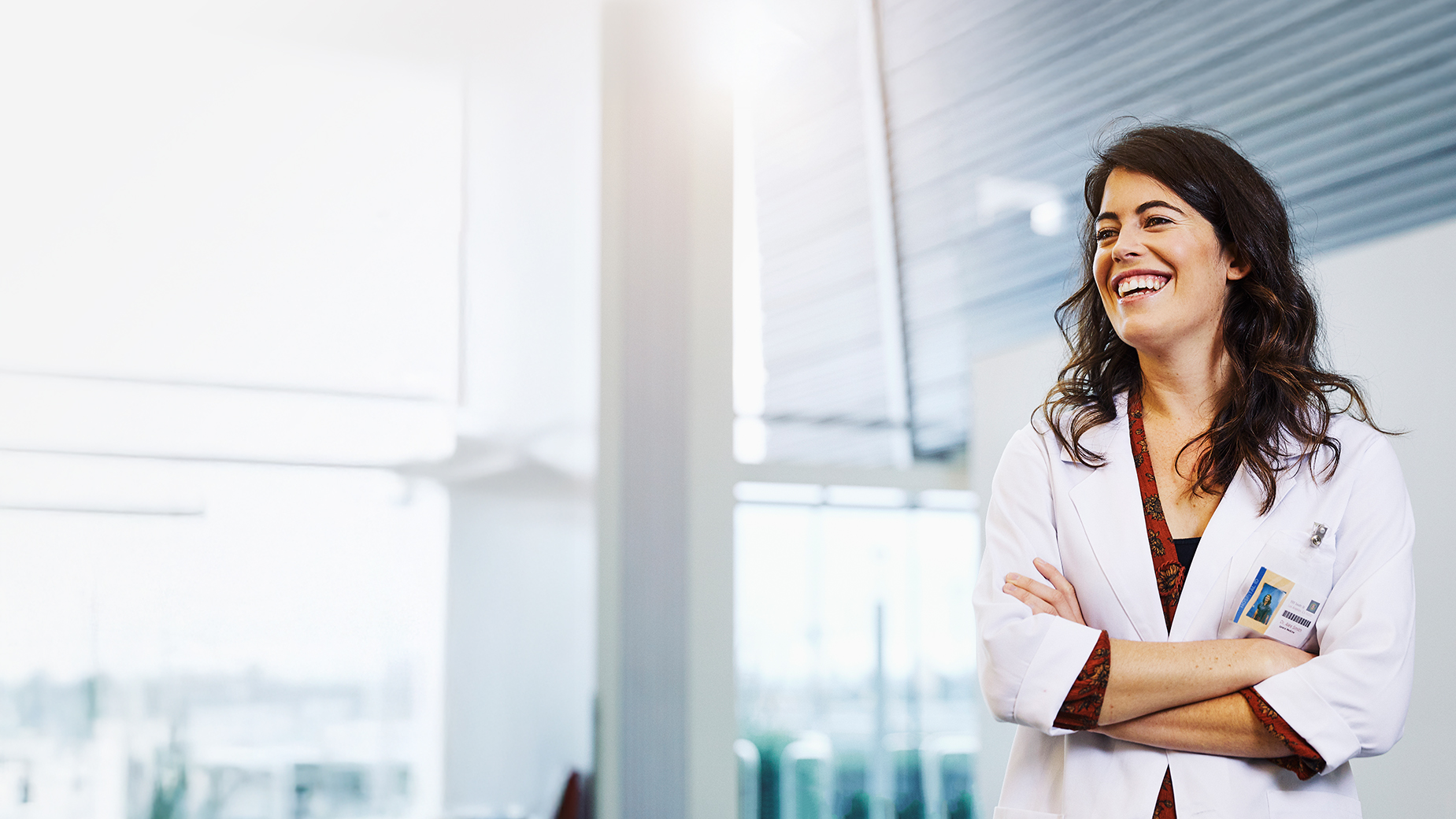 Woman in white lab coat smiling with arms crossed, standing in a modern medical or clinical setting