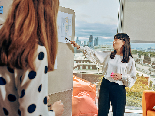 Two women in a modern and light office; one is pointing at a wireframe on the whiteboard, while the other is listening. A cityscape is visible through large windows.