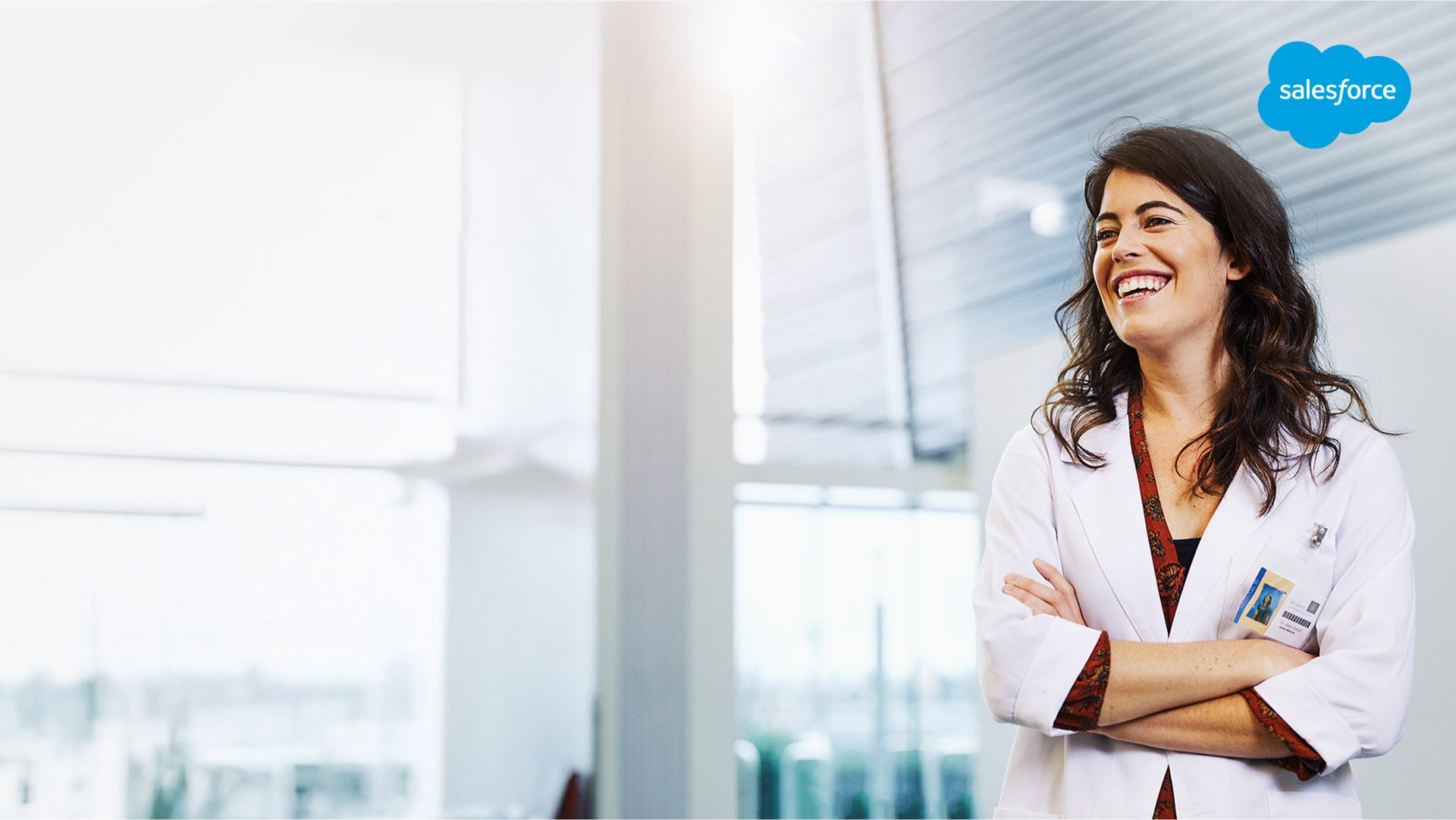 Woman in white lab coat smiling with arms crossed, standing in a modern medical or clinical setting