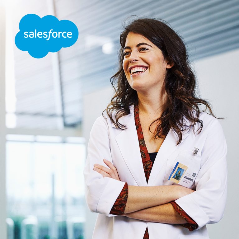 Woman in white lab coat smiling with arms crossed, standing in a modern medical or clinical setting