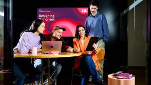 IBM iX Team arbeitet gemeinsam in einem modernen digitalen Studio mit Laptops und Tablets vor einem ‚Designing Tomorrow‘‑Screen und repräsentiert unseren human‑zentrierten Ansatz im Digital Experience Design.