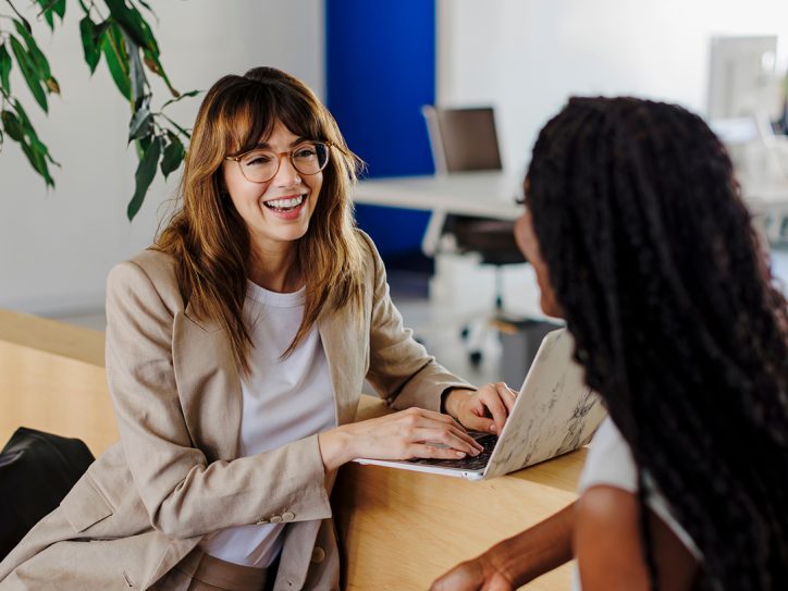 Zwei Frauen unterhalten sich lächelnd in einem modernen Büro, eine arbeitet an einem Laptop.